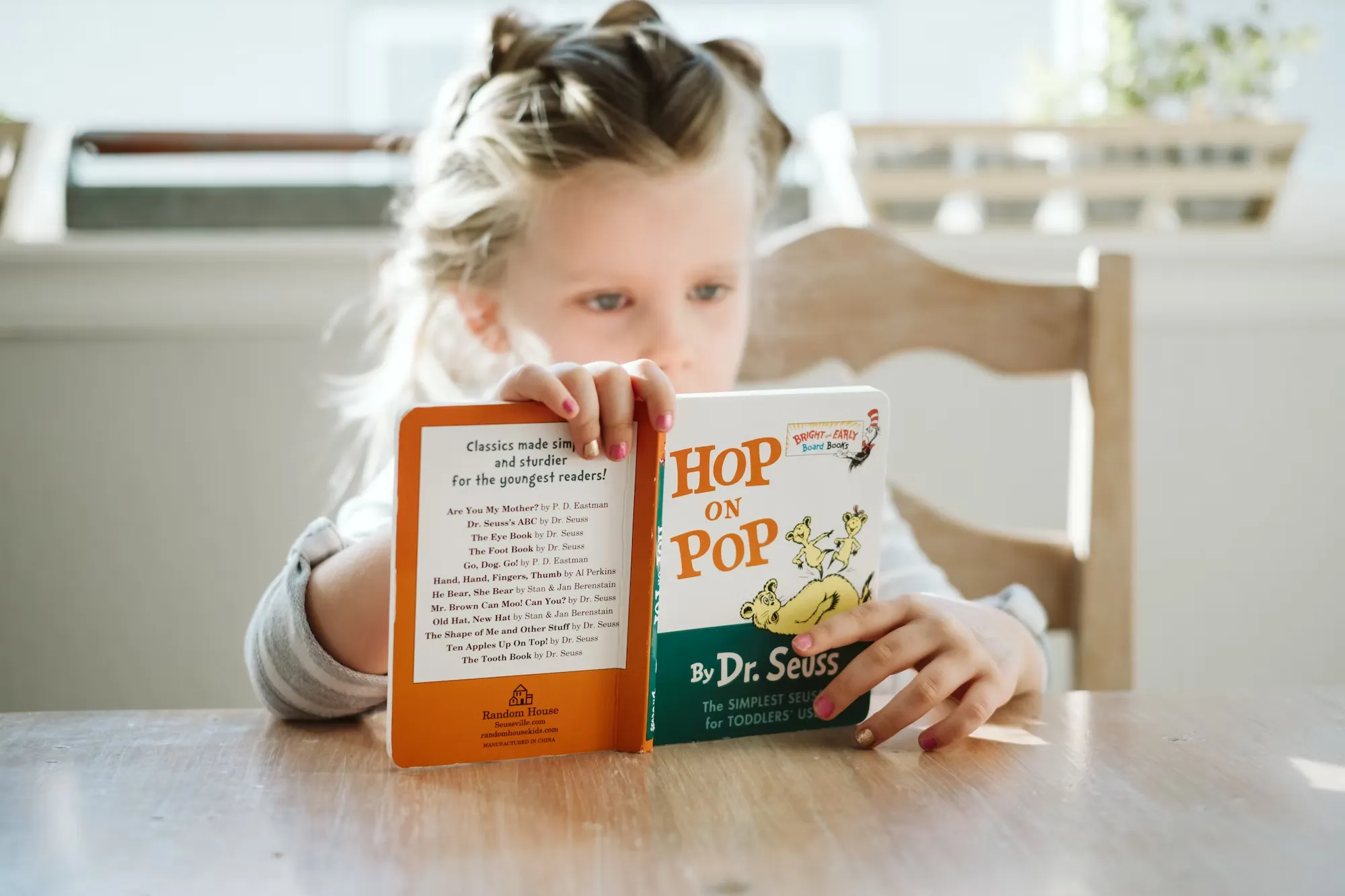 

The image shows a young girl, wearing glasses and a backpack, sitting on a chair and reading a book while using a tablet computer. She is smiling and looks engaged in her reading. This image illustrates how technology has enabled children to access books